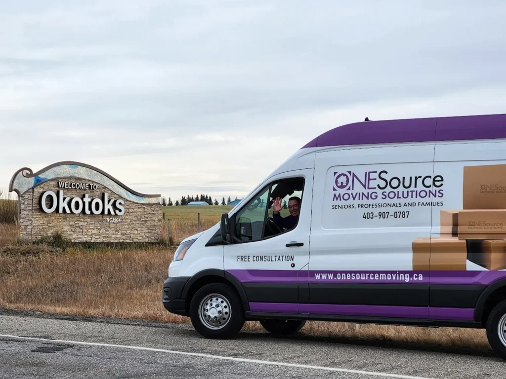 ONESource Moving Solutions van parked beside the Welcome to Okotoks town sign, illustrating senior downsizing and moving assistance in Okotoks.